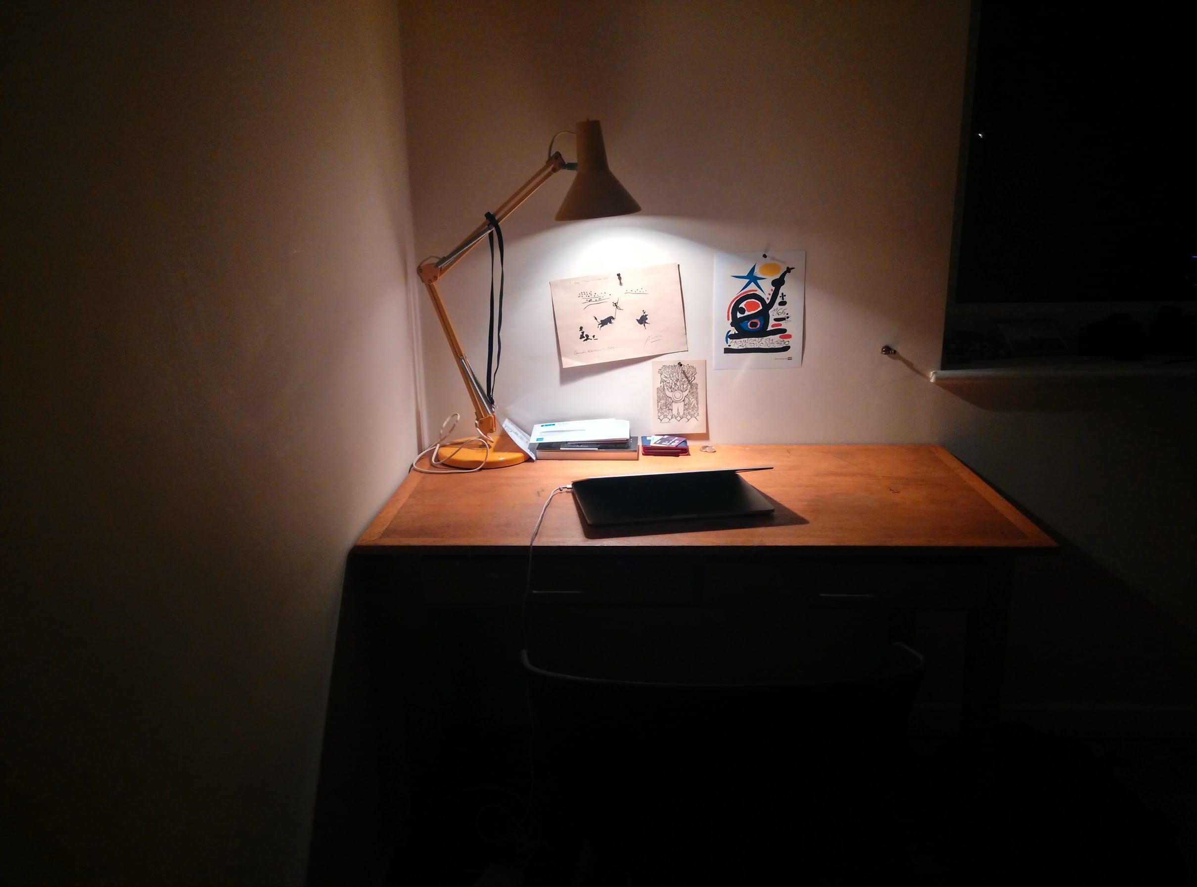 A wooden desk at night under a yellow anglepoise lamp, with prints pinned to the wall